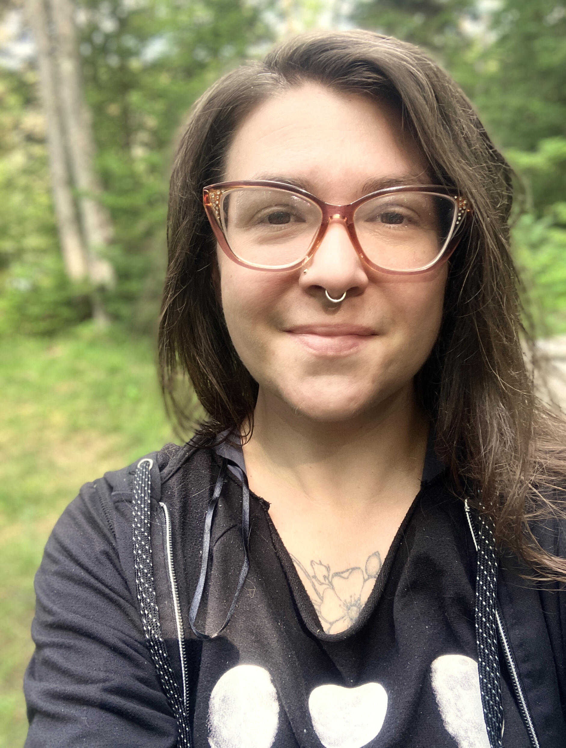 Image of a white woman smiling into the camera wearing glasses and a septum piercing, against a background of trees.